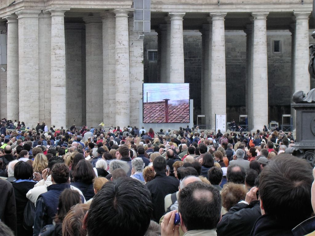 Fiéis se reúnem na Praça de São Pedro para acompanhar a escolha do novo papa — no telão, a fumaça branca surge no conclave de 2005 (Imagem: Tulumnes/CC-BY-SA-4.0)