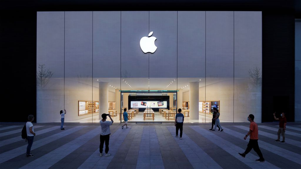 Queda de vendas obriga Apple a encerrar loja na China pela primeira vez 11 Fachada iluminada de uma Apple Store à noite, com várias pessoas observando a loja através das grandes janelas de vidro