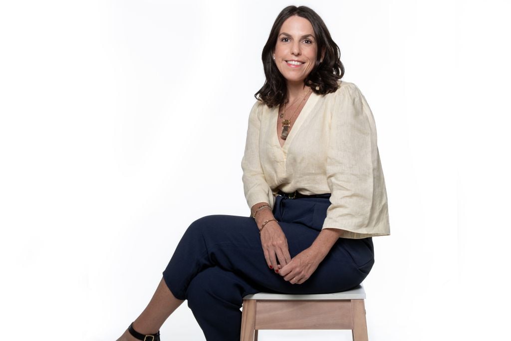 Diretora do Magalu Ads, Célia Goldstein, em uma foto com fundo branco, sentada em um banco, com blusa branca e saia preta