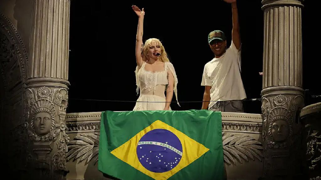 Lady Gaga usando vestido branco ao lado de um rapaz em cima de uma