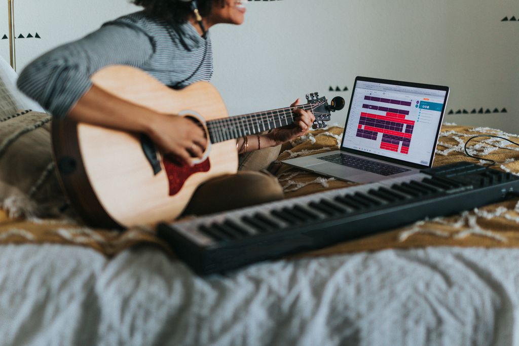 Mulher tocando violão e gravando a música pelo computador com um teclado musical ao lado