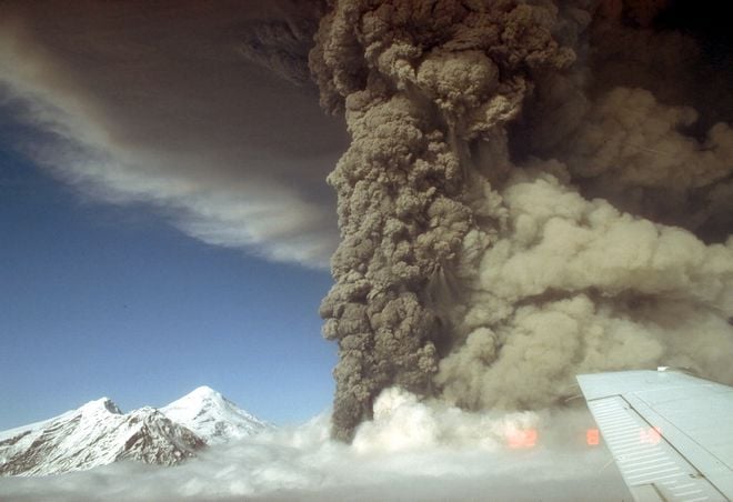 Foto de avião onde se vê parte da asa, montanhas e uma coluna de cinzas vulcânicas vindas da erupção do Pico da Cratera em 1992