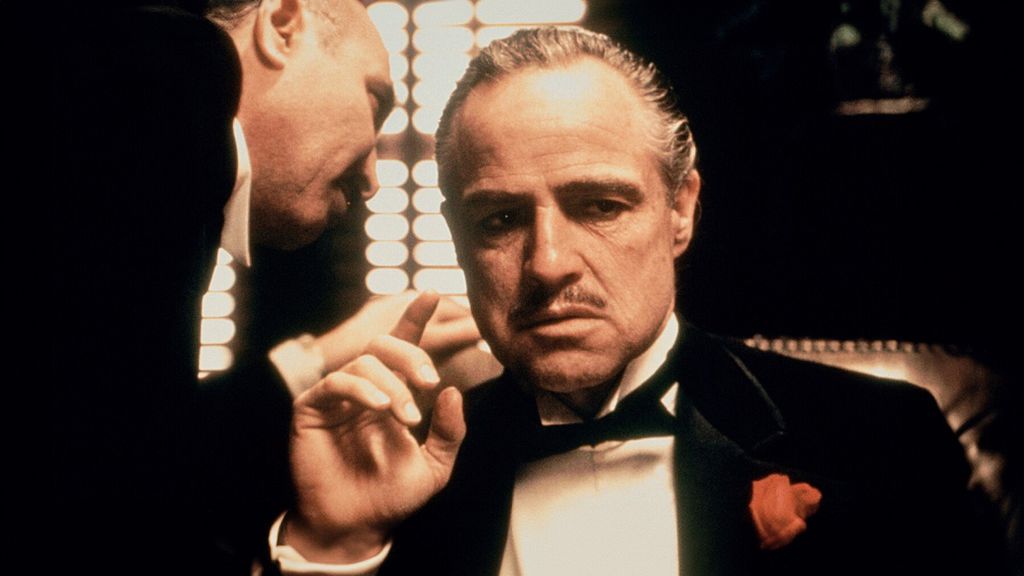Marlon Brando com um terno preto em O Poderoso Chefão escutando um homem falar próximo a seu ouvido.