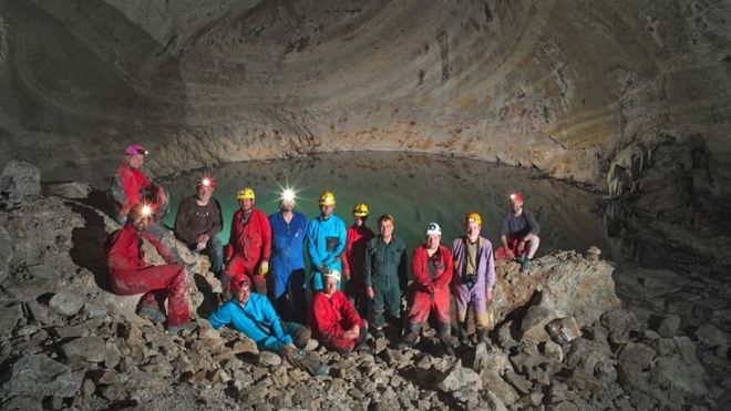 Pesquisadores com equipamentos de segurança posam em frente a uma caverna com um lago no fundo